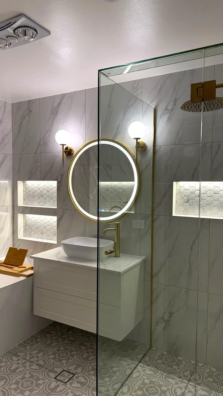 After: renovated bathroom with marble-look tiles, patterned floor, floating white vanity, round LED mirror and glass shower