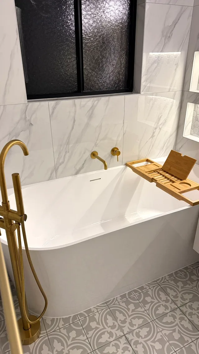 White bath with brass floor-mounted mixer and marble-look tiles under a black-framed window